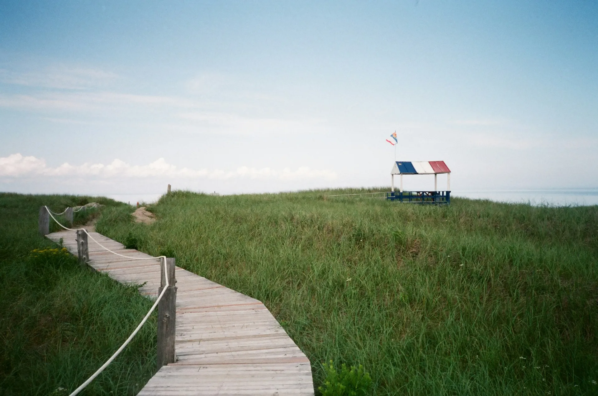 A boardwalk through dune grass toward a blue, white, and red lifeguard hut by the sea